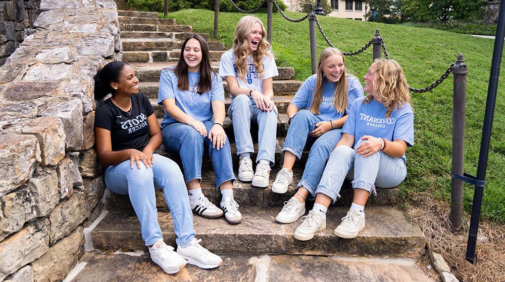 Five young women sitting on outdoor stone steps, wearing Covenant College apparel, laughing and talking together.